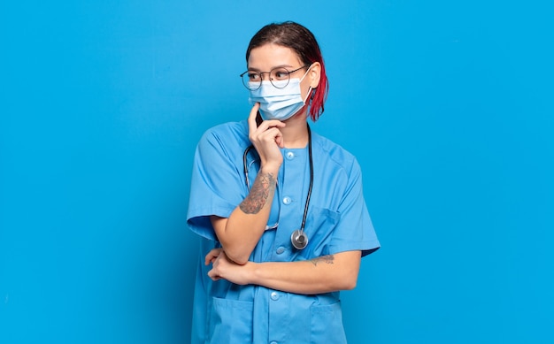 Mujer joven atractiva pelirroja sonriendo con una expresión feliz y segura con la mano en la barbilla, preguntándose y mirando hacia un lado. concepto de enfermera del hospital