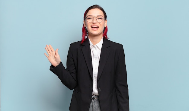 Mujer joven atractiva pelirroja sonriendo feliz y alegremente, saludando con la mano, dándole la bienvenida y saludándolo, o despidiéndose