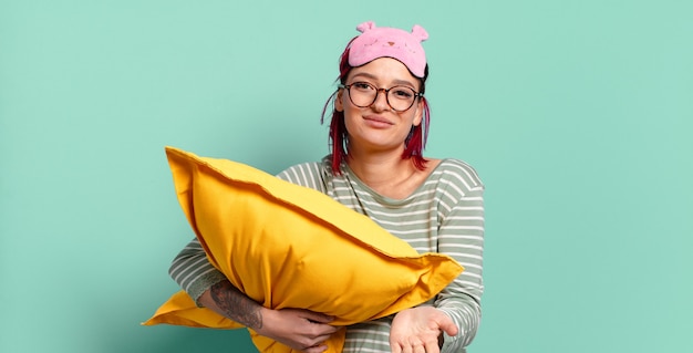 Mujer joven atractiva pelirroja sonriendo felizmente con mirada amistosa, segura y positiva, ofreciendo y mostrando un objeto o concepto y vistiendo pijamas.