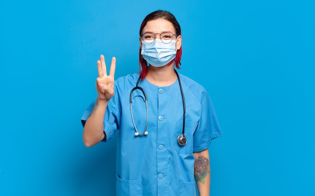 Mujer joven atractiva pelirroja sonriendo y mirando amigable, mostrando el número tres o tercero con la mano hacia adelante, contando hacia atrás. concepto de enfermera del hospital