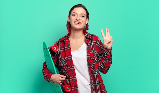 Foto mujer joven atractiva pelirroja sonriendo y mirando amigable, mostrando el número tres o tercero con la mano hacia adelante, contando hacia atrás y sosteniendo una patineta