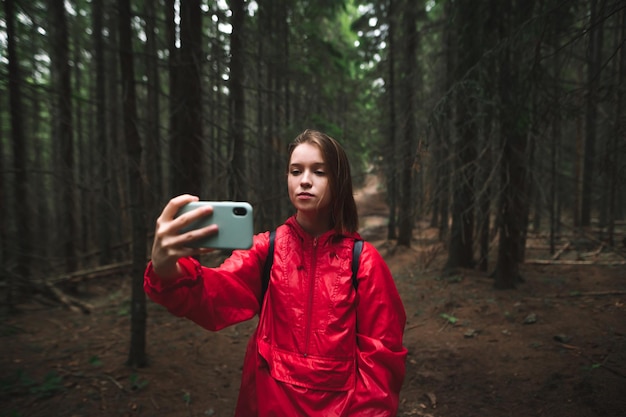 Una mujer joven atractiva posando para una foto selfie con un teléfono móvil en el bosque de abetos de montaña Una mujer turista con una chaqueta roja está tomando fotos en su teléfono inteligente mientras viaja al bosque