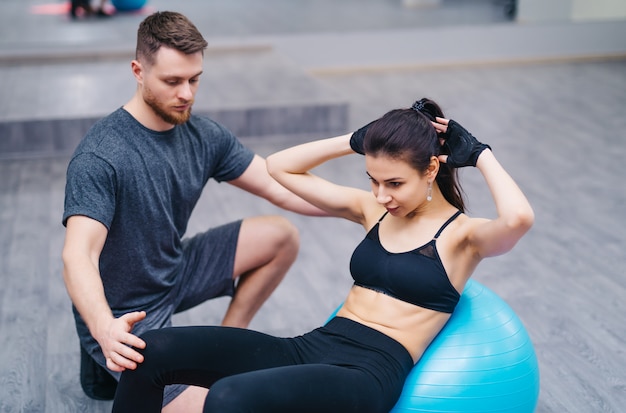 Mujer joven atractiva que hace los abdominales en una bola de la aptitud con el entrenador personal en el piso en gimnasio.