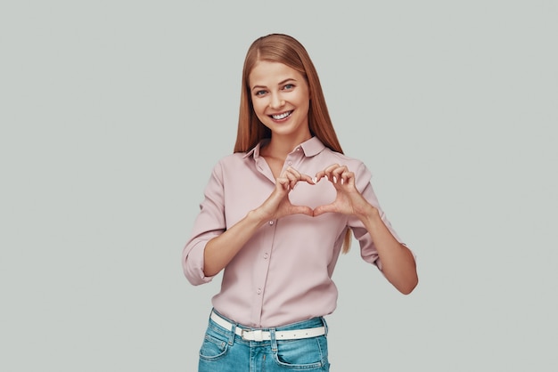 Mujer joven atractiva que muestra el corazón con las manos y sonriendo mientras está de pie contra el fondo gris