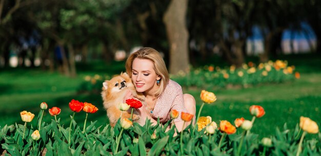 Mujer joven atractiva que sostiene el perro de Pomerania del perro afuera y que sonríe en la cámara, caminando en el parque.