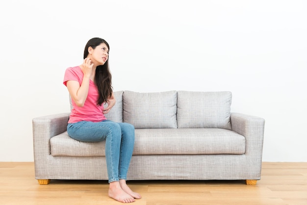 mujer joven y atractiva que tiene problemas de alergia sintiendo picazón en el cuerpo sentada en el sofá y mirando el fondo blanco usando la mano rascándose para calmar en el suelo de madera.