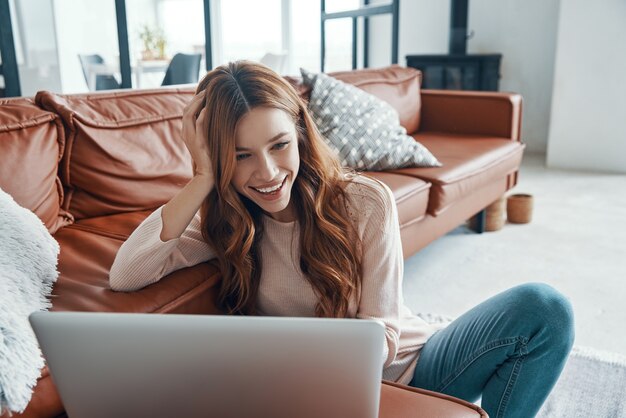 Mujer joven atractiva que trabaja en la computadora portátil y sonriendo mientras pasa tiempo en casa