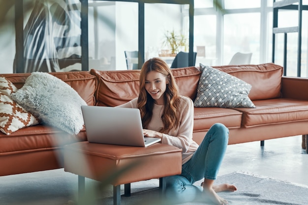 Mujer joven atractiva que trabaja en la computadora portátil y sonriendo mientras pasa tiempo en casa