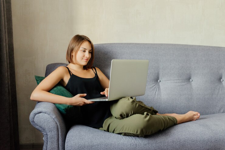 Mujer joven atractiva que usa una computadora portátil, mientras que miente en el sofá acogedor en casa. Tiempo de relajación.