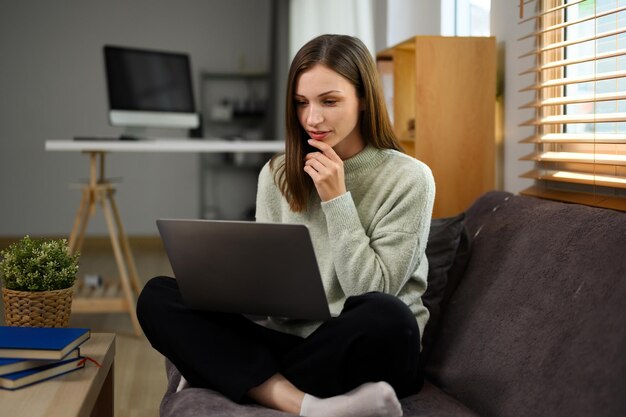 Mujer joven atractiva con ropa casual demandando a una computadora portátil sentada en el sofá en casa