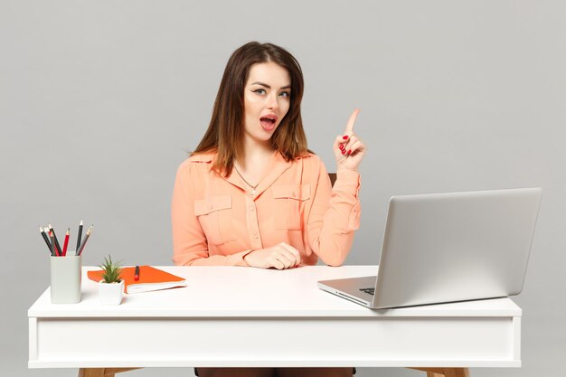 Una mujer joven y atractiva con ropa informal en colores pastel apuntando con el dedo índice hacia arriba se sienta a trabajar en el escritorio con una computadora portátil aislada en un fondo gris. Concepto de estilo de vida de carrera empresarial de logro. Simulacros de espacio de copia.