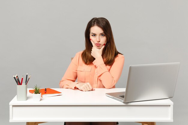 Una mujer joven y atractiva con ropa informal en colores pastel se puso la mano en la barbilla y se sentó en el escritorio con una computadora portátil aislada en un fondo gris. Concepto de estilo de vida de carrera empresarial de logro. Simulacros de espacio de copia.