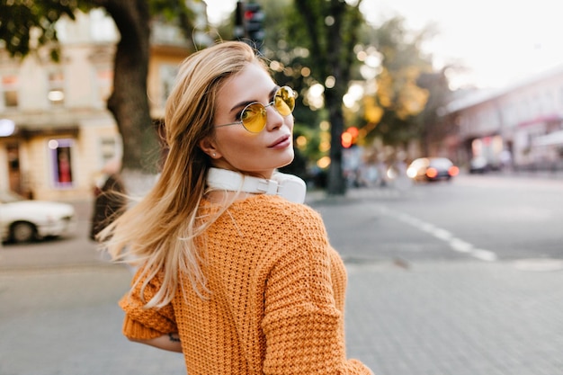 Mujer joven atractiva en suéter naranja de punto mirando por encima del hombro con una sonrisa suave. Hermosa chica rubia con gafas redondas y auriculares grandes posando al aire libre sobre fondo borroso.