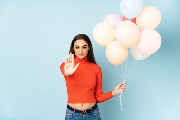 Foto mujer joven atrapar muchos globos aislados en la pared azul haciendo gesto de parada con su mano