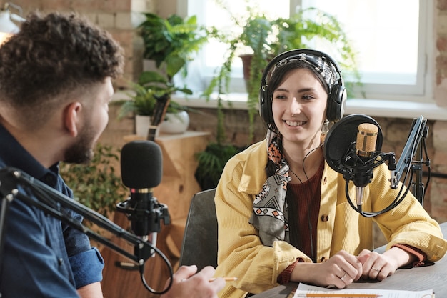 Mujer joven en auriculares dando una entrevista a un joven durante la transmisión en un estudio de radio