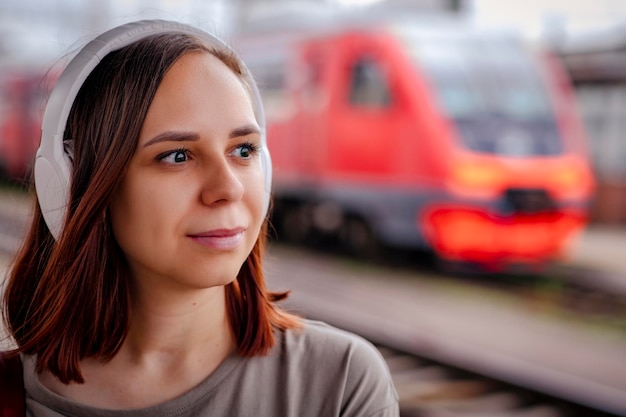 Mujer joven en auriculares inalámbricos se encuentra en la plataforma esperando el tren Retrato de una pasajera escuchando música en el fondo del tren borroso