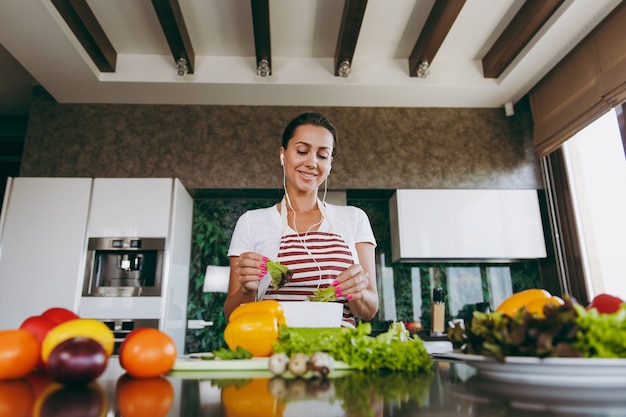 La mujer joven con auriculares en los oídos con verduras en las manos en la cocina con el portátil sobre la mesa