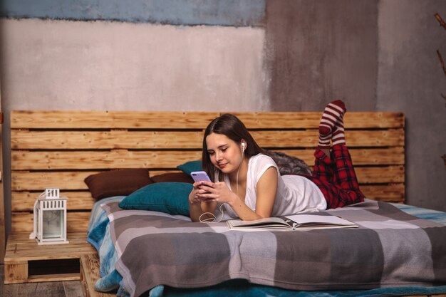 Una mujer joven con auriculares y un teléfono móvil en sus manos escucha las lecciones y mira un libro mientras está acostada en la cama. Formación en línea, ocio.