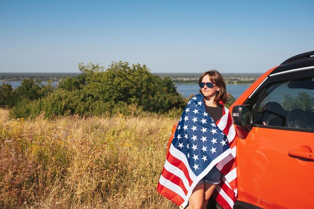 Mujer joven con la bandera de los Estados Unidos de América parada cerca del auto