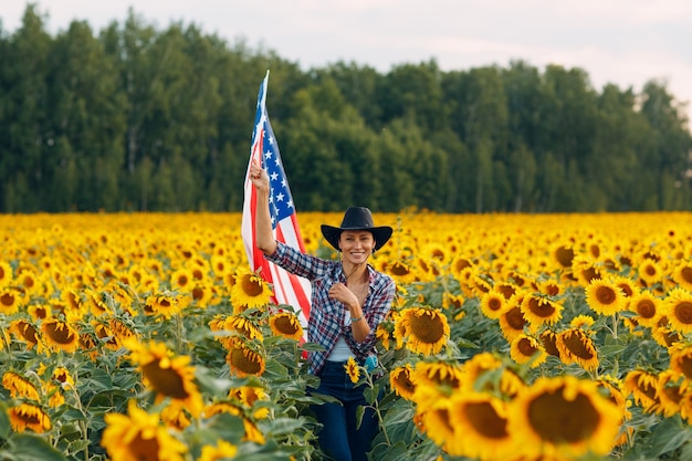 Mujer joven con bandera estadounidense en el campo de girasol. 4 de julio concepto del día de la independencia de Estados Unidos.