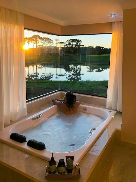 Mujer joven en la bañera de hidromasaje en el hotel con una vista de la naturaleza capturando la puesta de sol