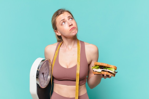 Foto mujer joven bastante deporte dudando o expresión incierta y sosteniendo una balanza y un sándwich