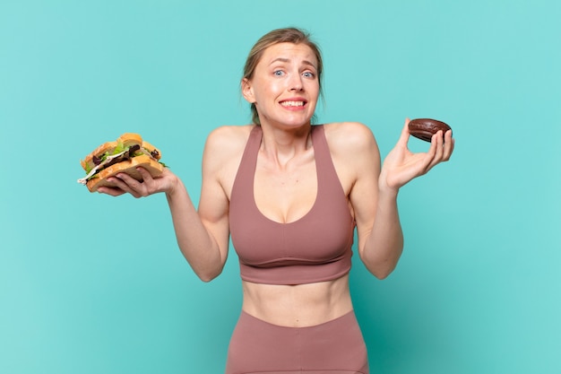 Mujer joven bastante deporte dudando o expresión incierta y sosteniendo un sándwich y una rosquilla