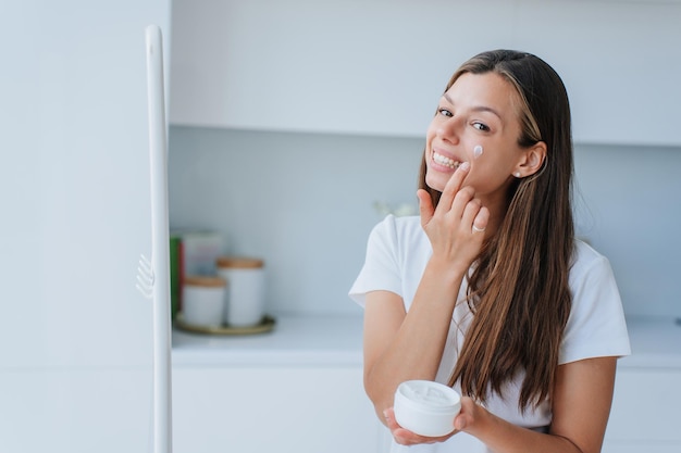 Mujer joven bastante italiana con el pelo largo y suelto de pie en la sala de estar pone crema para la piel en la cara amplias sonrisas satisfechas con productos para el cuidado de la piel y un estilo de vida saludable Cosméticos Mujer satisfecha en casa