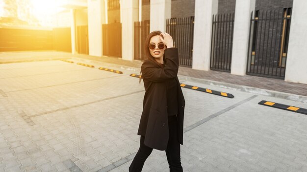 Mujer joven bastante moderna fresca estadounidense en ropa juvenil de moda con gafas de sol con estilo disfrutar de caminar al aire libre al atardecer. Modelo de moda hermosa chica de moda en traje negro vintage viaja en la calle.