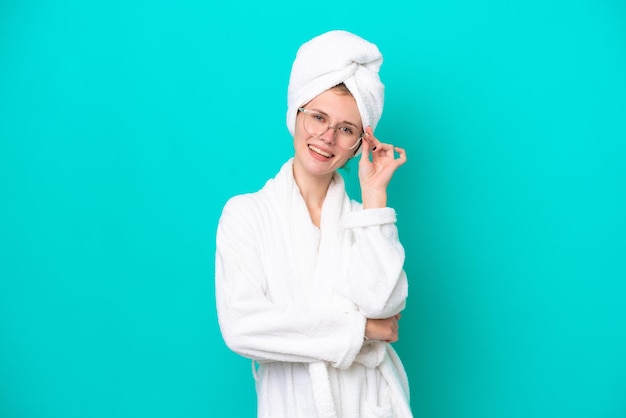 Mujer joven en una bata de baño aislada sobre fondo azul con gafas y feliz