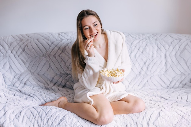 Mujer joven en bata de baño blanca con palomitas de maíz viendo películas en sof