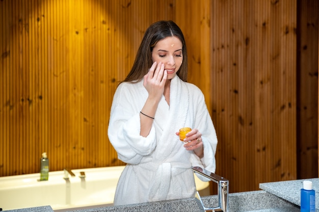 Mujer joven en bata de baño blanca de pie frente al espejo en el baño y aplicando crema cosmética en la cara durante la rutina diaria de cuidado de la piel