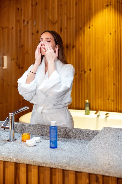 Mujer joven en bata de baño blanca de pie frente al espejo en el baño y aplicando crema cosmética en la cara durante la rutina diaria de cuidado de la piel