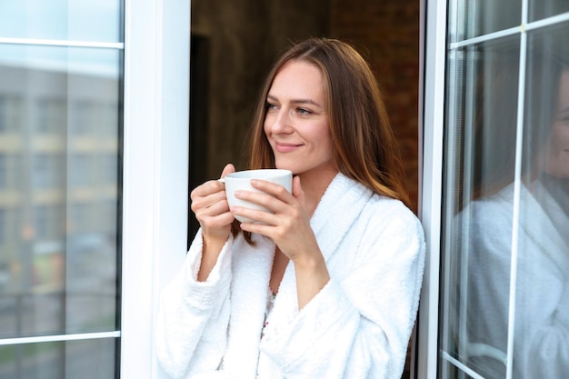 Una mujer joven con una bata de baño blanca sostiene una taza en sus manos. Descanse en el hotel.