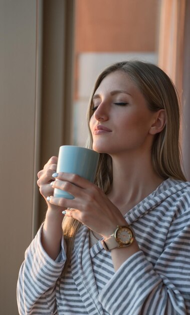 mujer joven en bata de baño disfrutando del café de la tarde y el hermoso paisaje de la puesta de sol de la ciudad mientras está de pie junto a la ventana