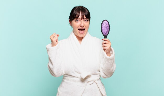 Mujer joven con bata de baño que se siente sorprendida, emocionada y feliz, riendo y celebrando el éxito, diciendo ¡guau!