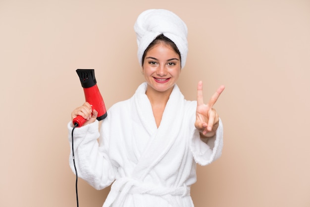 Mujer joven en una bata de baño con secador de pelo sonriendo y mostrando el signo de la victoria