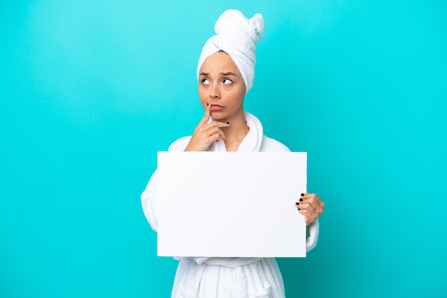 Mujer joven en bata de baño con toalla aislada de fondo azul sosteniendo un cartel vacío y pensando