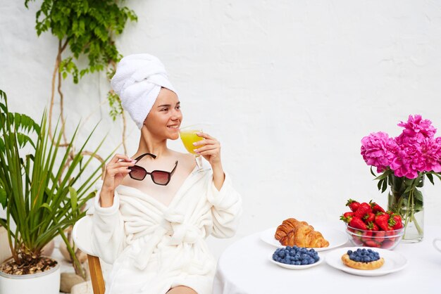 Una mujer joven en una bata de baño con una toalla en la cabeza disfruta de sus vacaciones en el resort desayuna café con un croissant y fresas