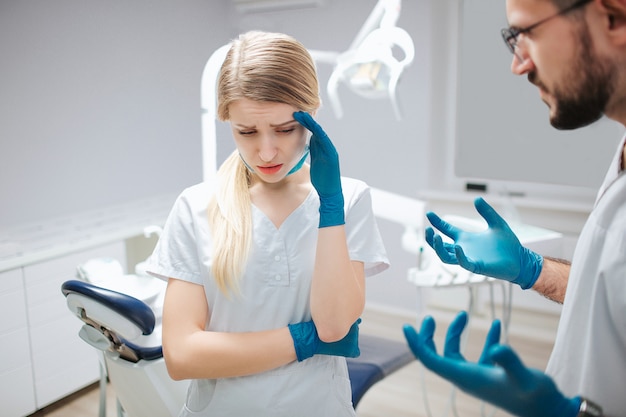 Mujer joven en bata blanca de pie en la habitación con el dentista. Ella se toma de la mano y sufre de dolor de cabeza. El hombre mira a la mujer y preocupado.