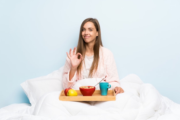 Mujer joven en bata con desayuno mostrando un signo bien con los dedos