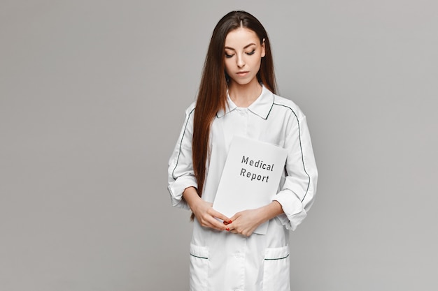 Mujer joven en bata médica blanca posando con un informe médico en sus manos en el fondo gris. Copie espacio para su texto y producto. Concepto de salud. Modelo femenino en la imagen de un médico