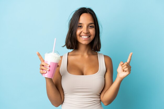 Mujer joven con batido de fresa aislado en la pared azul apuntando hacia una gran idea