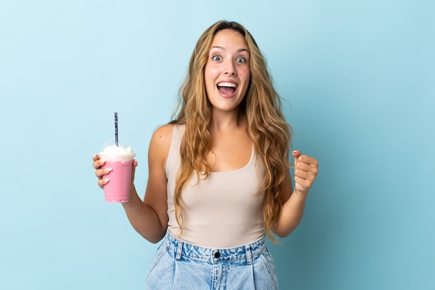 Mujer joven con batido de fresa aislado en la pared azul celebrando una victoria en la posición ganadora