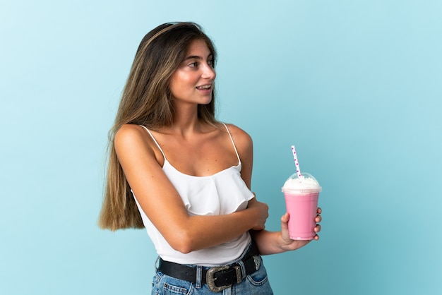 Mujer joven con batido de fresa aislado en la pared azul mirando hacia el lado y sonriendo