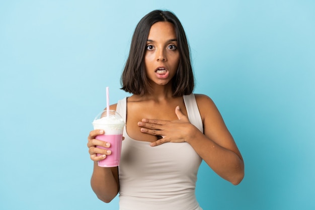 Mujer joven con batido de fresa aislado en la pared azul sorprendido y conmocionado mientras mira a la derecha