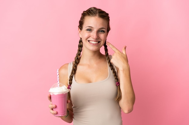 Mujer joven con batido de fresa aislado en rosa dando un pulgar hacia arriba gesto