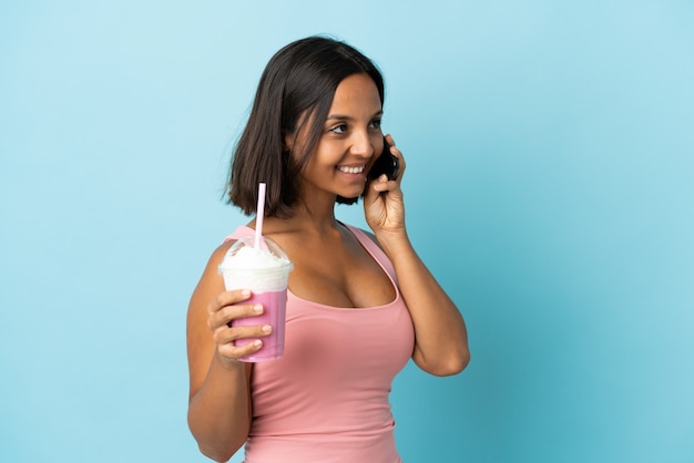 Mujer joven con batido de fresa aislado sobre fondo azul manteniendo una conversación con el teléfono móvil