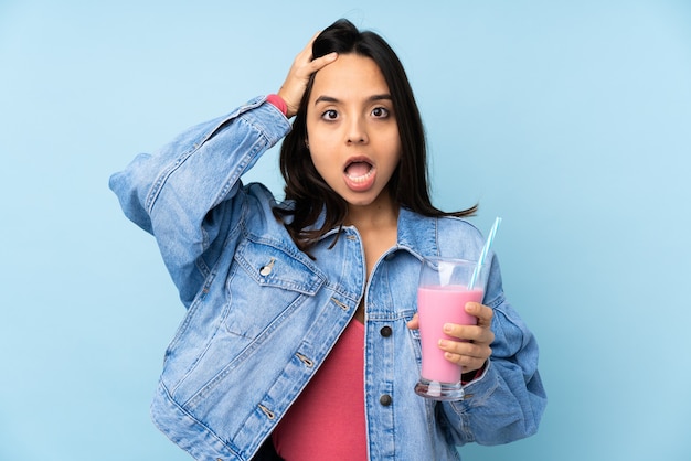 Mujer joven con batido de fresa en azul aislado con expresión de sorpresa