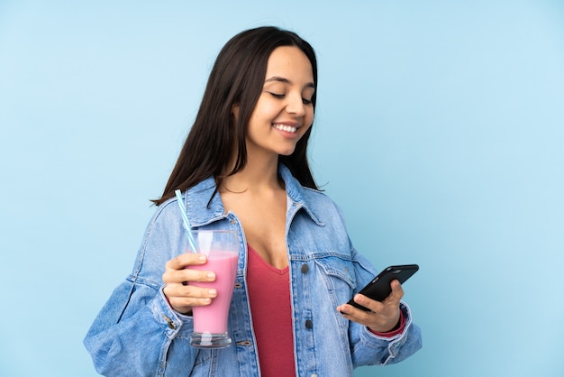 Mujer joven con batido de fresa en azul aislado sosteniendo café para llevar y un móvil